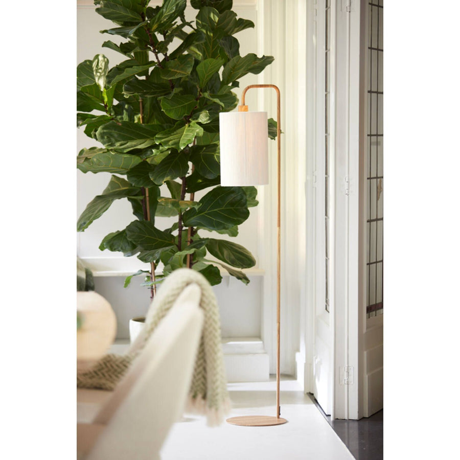 Een gouden staande lamp met een witte lampenkap staat naast een grote potplant en een witte bank met een groene deken.