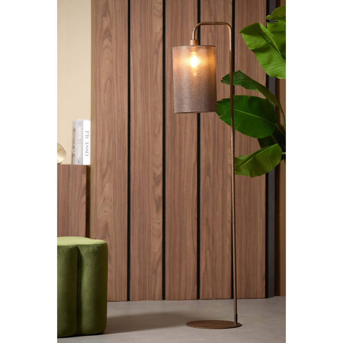 Een staande lamp met een kap met structuur staat naast een plant en een houten muur.
