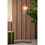 Een staande lamp met een kap met structuur staat naast een plant en een houten muur.