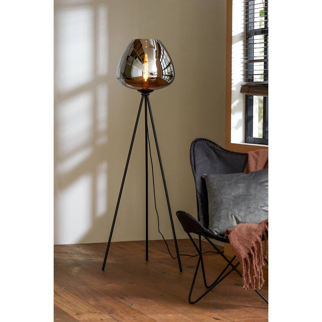 Zwarte staande lamp op een statief met een amberkleurige glazen kap naast een stoel en een raam.