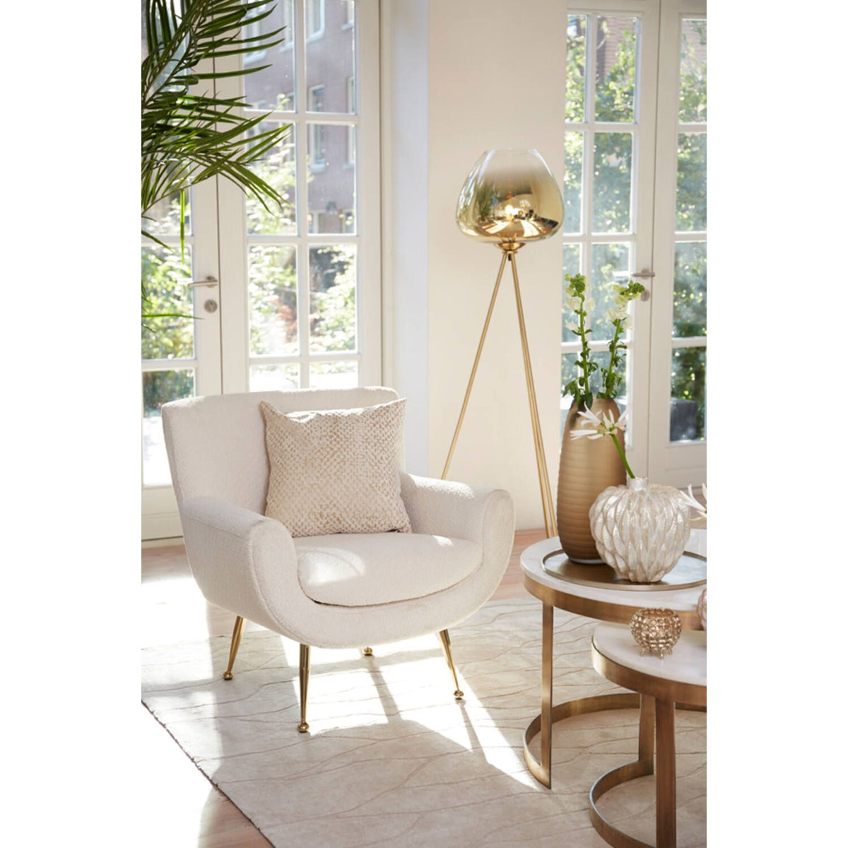Gezellige woonkamer met crèmekleurige fauteuil, gouden driepootlamp, marmeren salontafels en planten bij het raam.