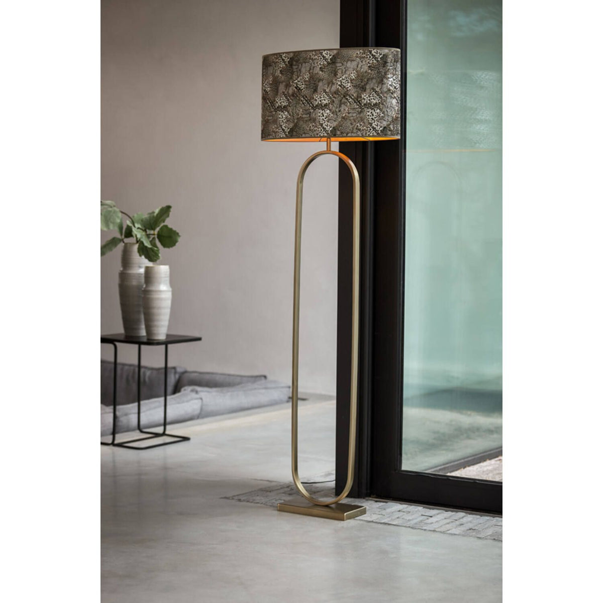 Gouden staande lamp met een lampenkap met patroon, naast een zwarte tafel met een glazen deur.