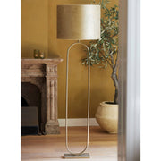 Gouden staande lamp met ovale rand en gouden kap naast een olijfboom in een kamer met een open haard.